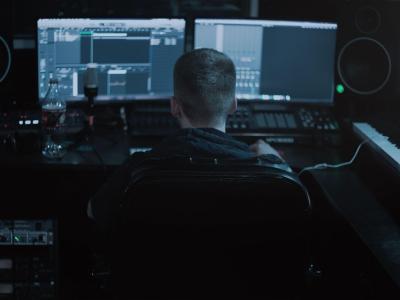 Man seated in a dark workstation surrounded by monitors and other electronic music production equipment